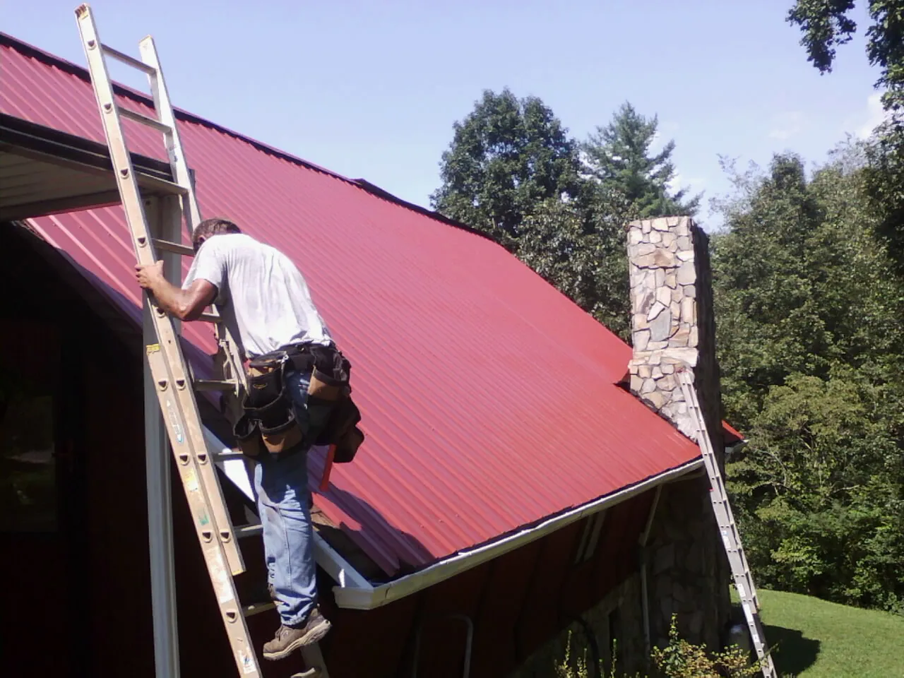 Roofer on a ladder beside a red metal roof and stone chimney — Soffit & Fascia Repair by Roofing Brothers in Aumsville